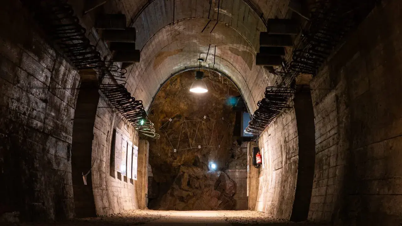 Mroczny tunel, idealny na riese zwiedzanie. Widać odsłonięte skały, metalowe elementy konstrukcyjne i tablice informacyjne.