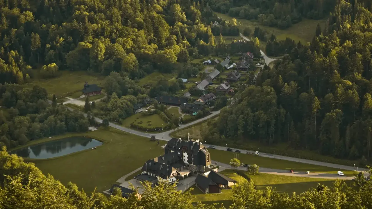 Widok z góry na osadę w lesie, przypominającą legendarną **Wilczą Górę**. Domy i hotel otoczone zielenią, z małym stawem.