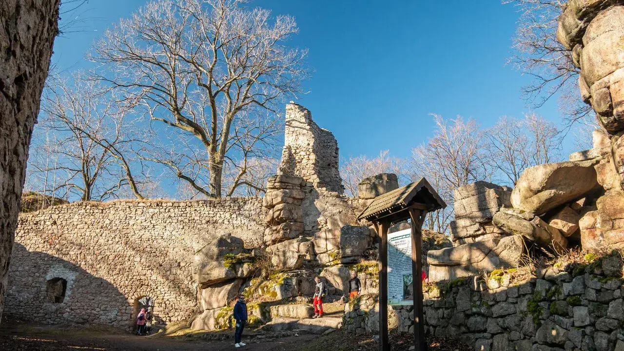 Ruiny zamku Bolczów w słoneczny dzień. Kamienne mury, skalne formacje i nagie drzewa tworzą malowniczy krajobraz.