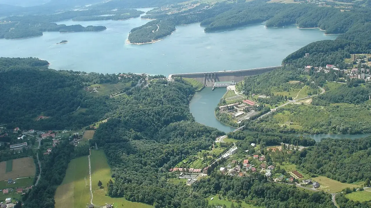 Widok z lotu ptaka na zapora wodna w Myczkowcach, otoczona zielonymi wzgórzami i rozległym jeziorem.