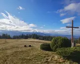 Krawcowy Wierch wita słonecznym niebem i chmurami. Drewniany krzyż góruje nad polaną, a w oddali widać pasmo górskie.