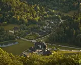 Widok z góry na osadę w lesie, przypominającą legendarną **Wilczą Górę**. Domy i hotel otoczone zielenią, z małym stawem.