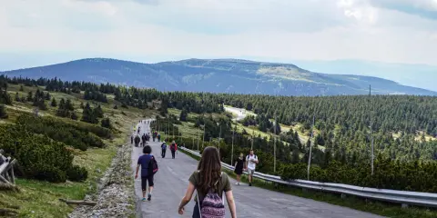 Grupa turystów wędruje pod górę w kierunku pradziad szczyt. W oddali widać pasma górskie i lasy.