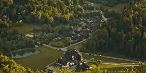 Widok z góry na osadę w lesie, przypominającą legendarną **Wilczą Górę**. Domy i hotel otoczone zielenią, z małym stawem.