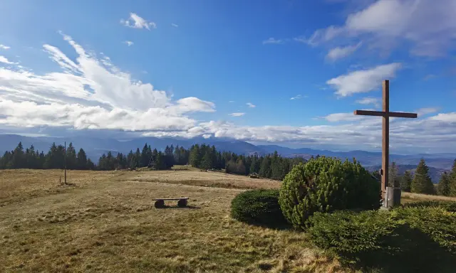 Krawcowy Wierch wita słonecznym niebem i chmurami. Drewniany krzyż góruje nad polaną, a w oddali widać pasmo górskie.