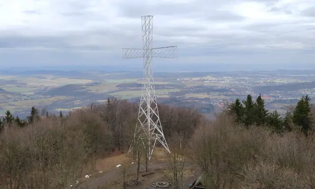 Wielki metalowy krzyż góruje nad lasem na Chełmcu. W dole rozciąga się panorama miasta i pagórkowatego krajobrazu.