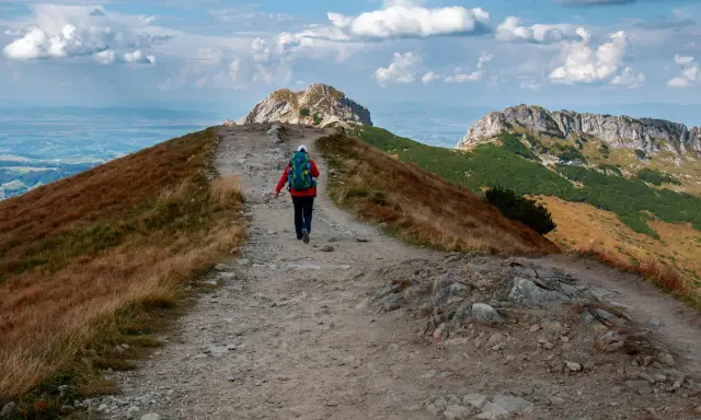 Wędrowiec na szlaku w **Czerwonych Wierchach**. Kamienista ścieżka prowadzi przez trawiaste zbocza ku skalistym szczytom.