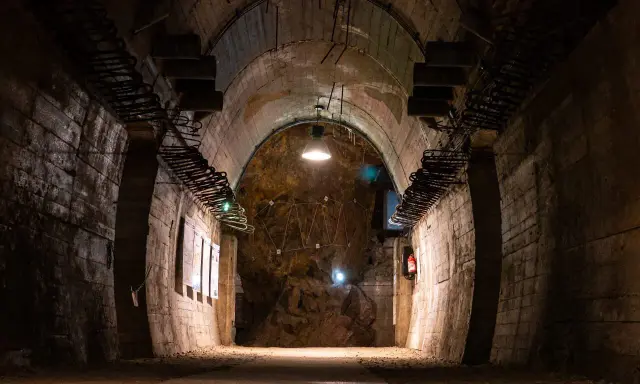 Mroczny tunel, idealny na riese zwiedzanie. Widać odsłonięte skały, metalowe elementy konstrukcyjne i tablice informacyjne.