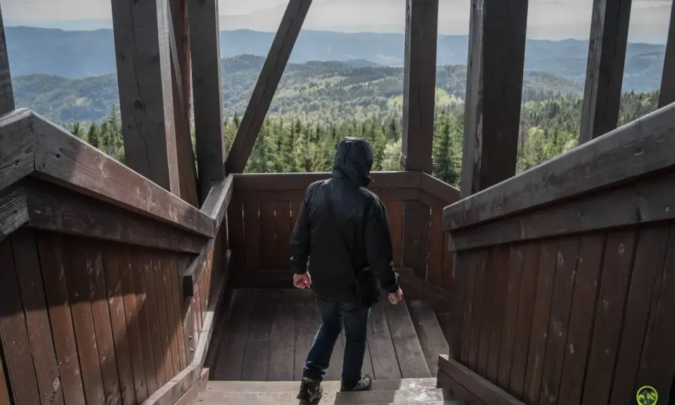 Osoba w czarnej kurtce schodzi po drewnianych schodach wieży widokowej. W oddali rozciąga się malowniczy krajobraz gór i lasów, który zapiera dech w piersiach.