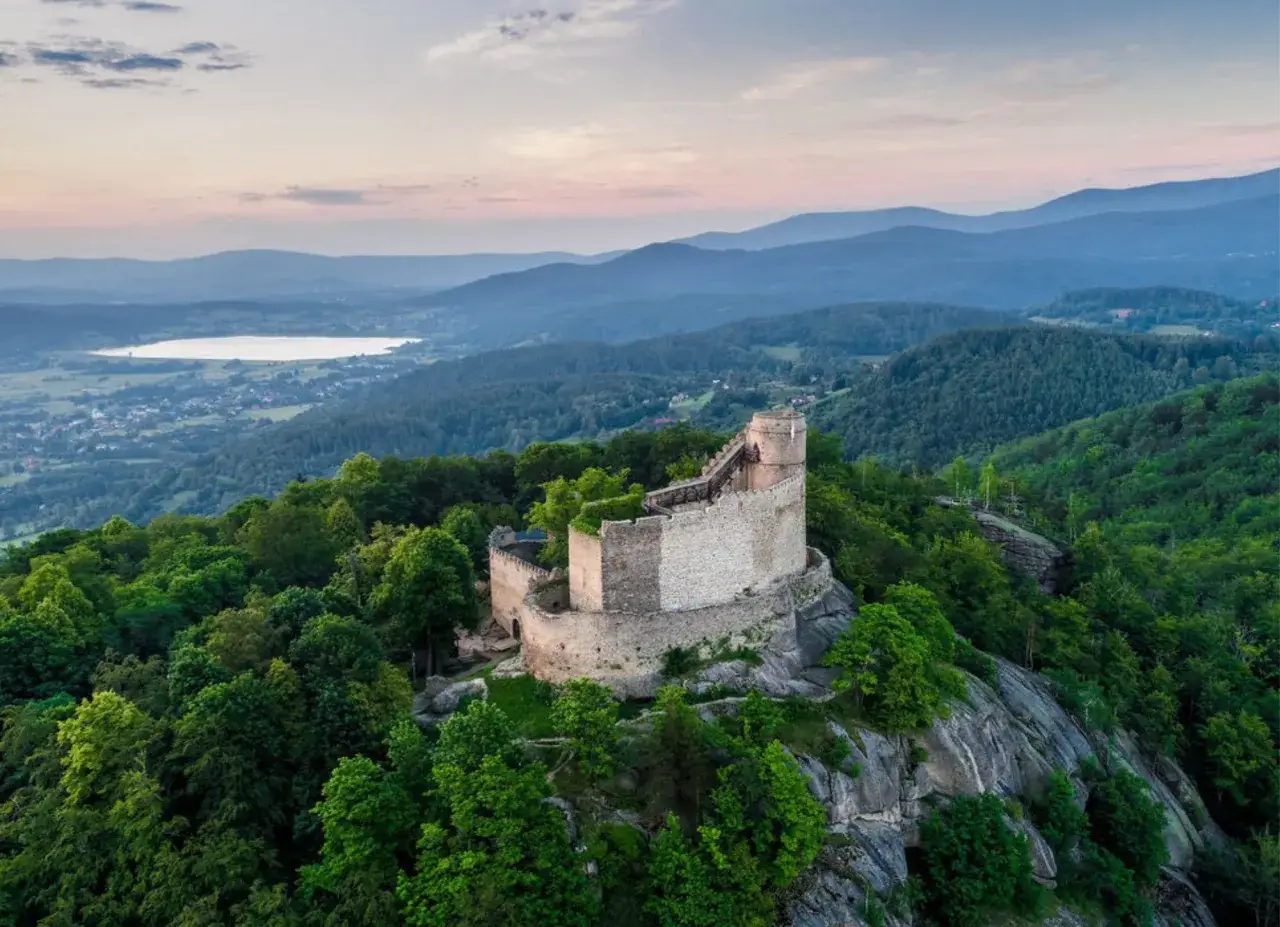 Zamek Chojnik, majestatyczna twierdza na skale, otoczona zielonymi lasami i górami, z jeziorem w oddali.